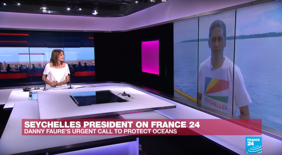 Woman wearing white sitting at desk in television broadcast facilities studio looking at screen on wall with image of seychelles president danny faure reporting live from indian ocean
