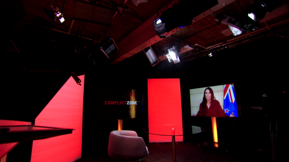 Rows of ceiling lights above chair and table in front of red light panels and video monitor showing image of woman in london content studio