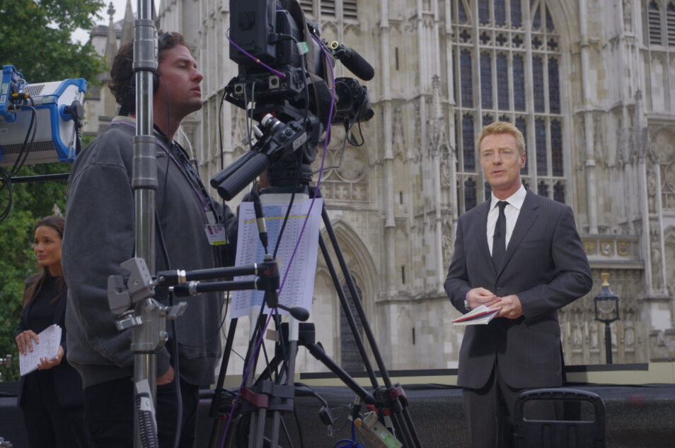 Man wearing a suit and black tie standing in front of westminster abbey and facing a man operating a video camera while broadcasting live at the state funeral of queen elizabeth ii
