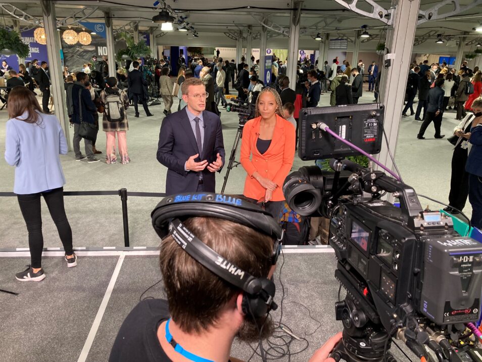 Man dressed in suit and tie and woman dressed in orange jacket standing in front of conference attendees and facing a video camera being operated by a man wearing headphones as part of broadcast services at un climate talks in Glasgow 2021
