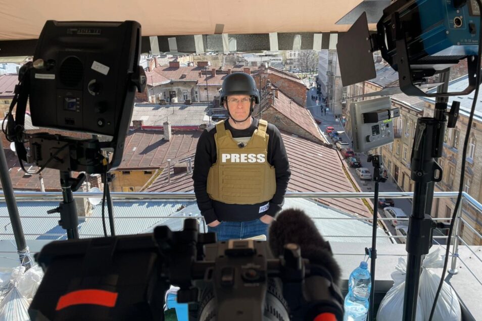 Live news reporter wearing helmet and flak jacket looking towards a video camera in front of rooftops of houses in ukraine