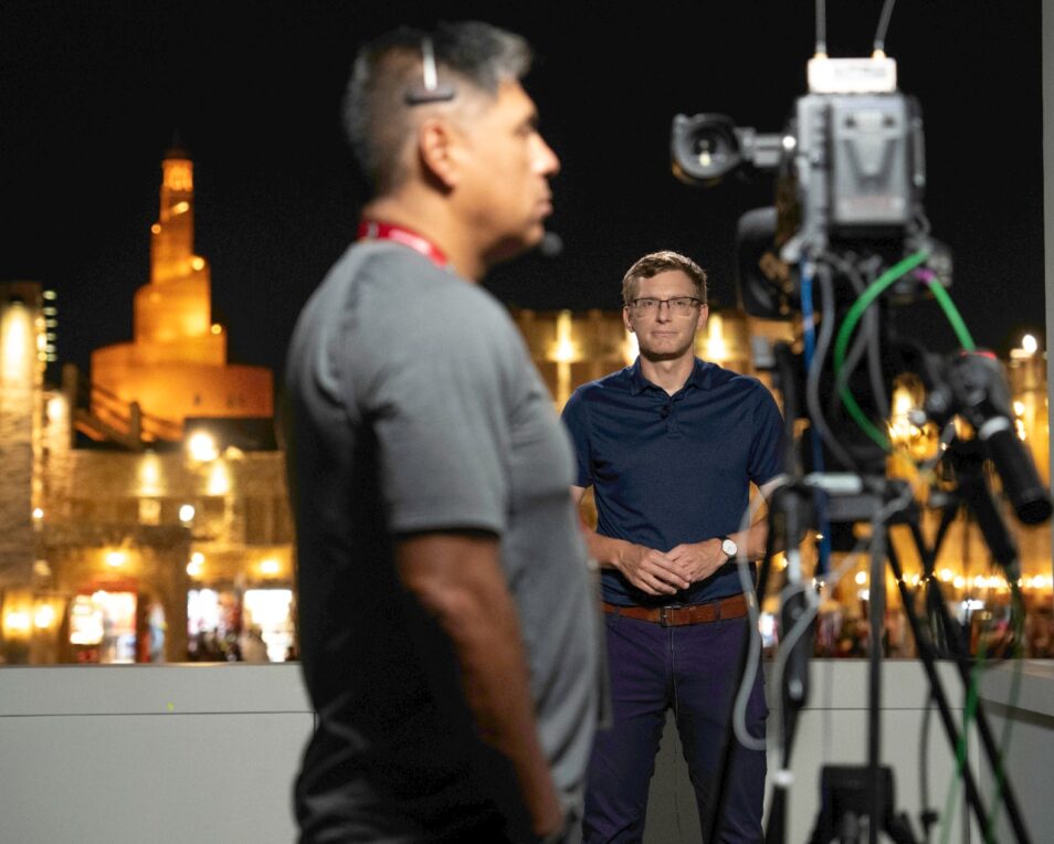 Live news reporter standing in front of floodlit buildings in doha looking towards a video camera and camera operator wearing a grey shirt