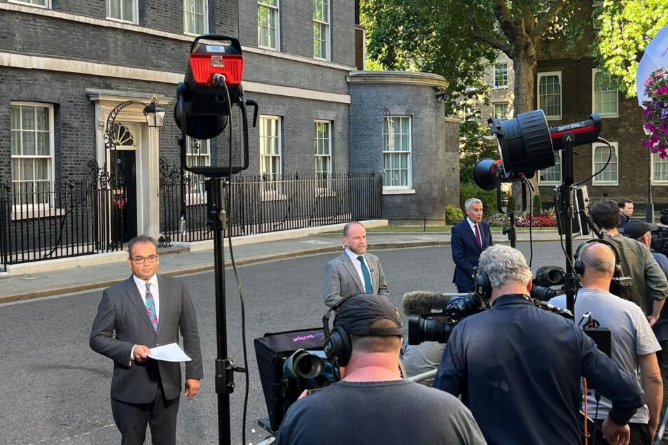 Three male journalists wearing suits and ties standing in downing street in front of a row of camera operators with video equipment and lights reporting live on uk politics