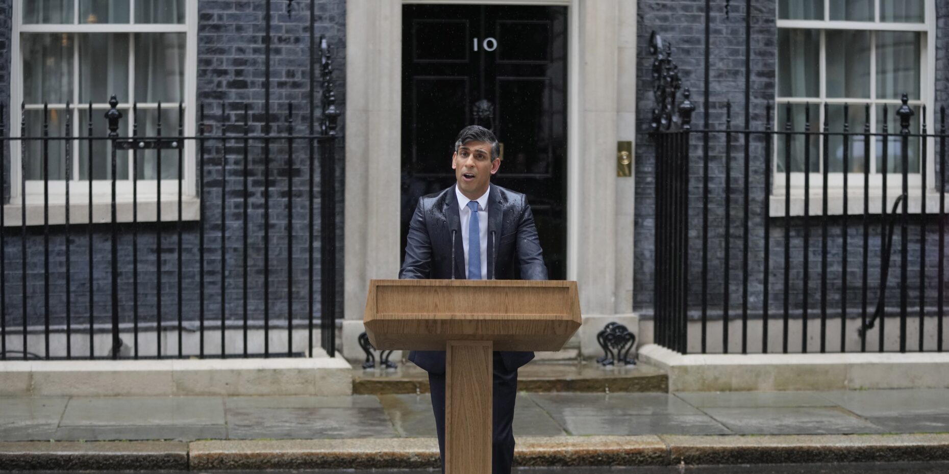 British prime minister rishi sunak wearing a suit and tie standing at a wooden lectern in front of the door to number 10 downing street announcing the 2024 uk general election